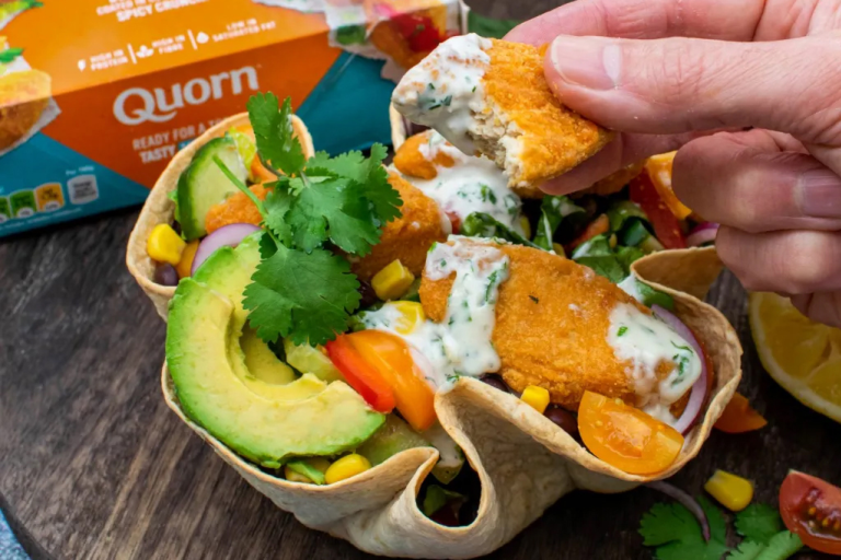A tortilla bowl, filled with salad and Quorn Buffalo Wings sits on a wooden chopping board. A hand leans into frame holding a half eaten crispy Buffalo Wing