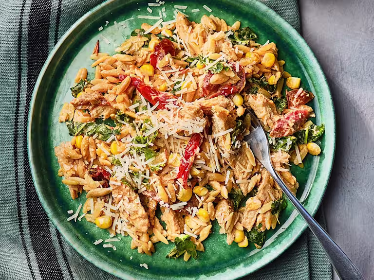 Orzo pasta in a creamy sauce with Quorn Vegetarian Fillets, sundried tomatoes, sweetcorn, kale, and parmesan cheese with a fork on the side.