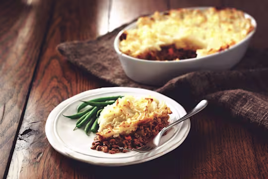 A white plate with a serving of sherpherd's pie and a side of green beans on top, with the baking dish of the whole pie in the background.