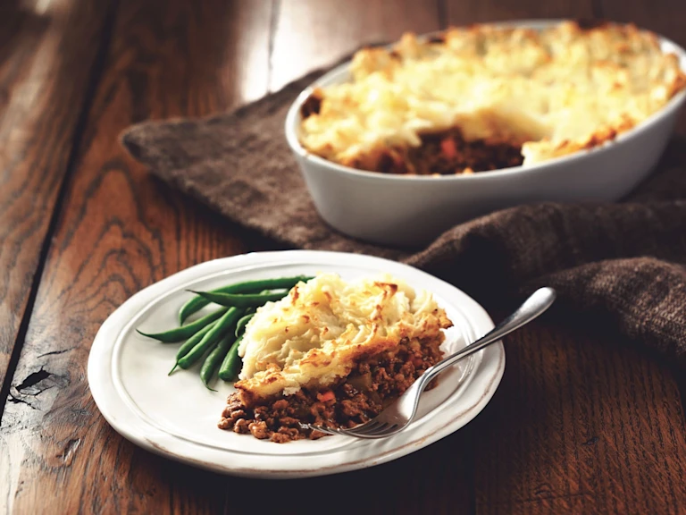 A white plate with a serving of sherpherd's pie and a side of green beans on top, with the baking dish of the whole pie in the background.