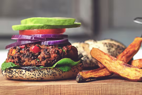 A veggie burger on a poppyseed bun topped with lettuce, tomato, red onions, and avocado with thick-cut fries on the side.