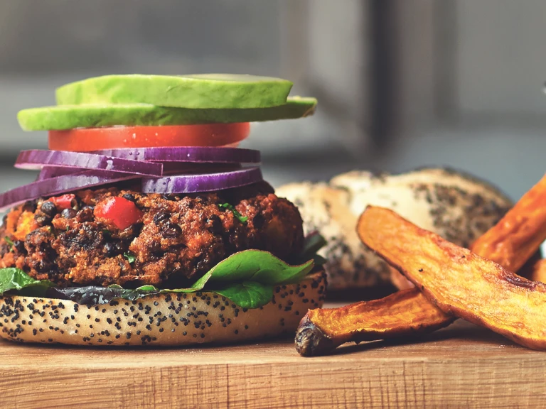 A veggie burger on a poppyseed bun topped with lettuce, tomato, red onions, and avocado with thick-cut fries on the side.