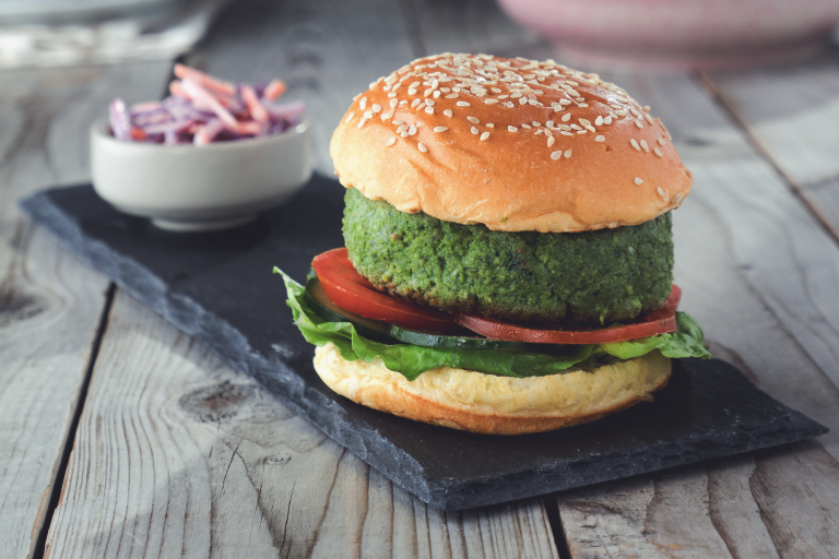 A burger made of Quorn Pieces and spinach on a sesame seed bun with sliced tomatoes, cucumber, and lettuce on a slate with a small dish of coleslaw in the background.