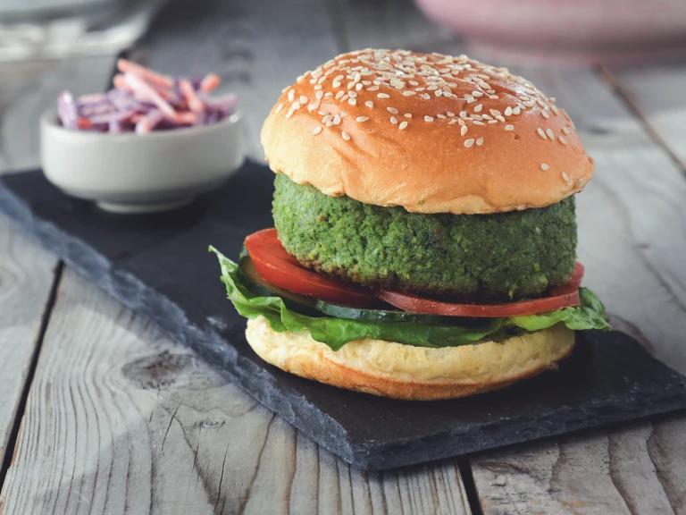 A burger made of Quorn Pieces and spinach on a sesame seed bun with sliced tomatoes, cucumber, and lettuce on a slate with a small dish of coleslaw in the background.