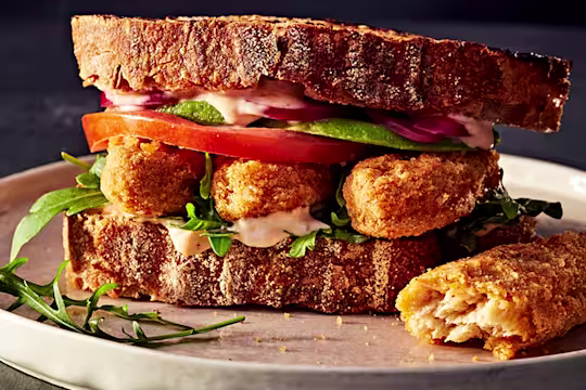 Crispy vegetarian chicken sandwich with tomato, onion and salad in sourdough bread served on a white plate with a cut Quorn Salt and Pepper Tender on the side.