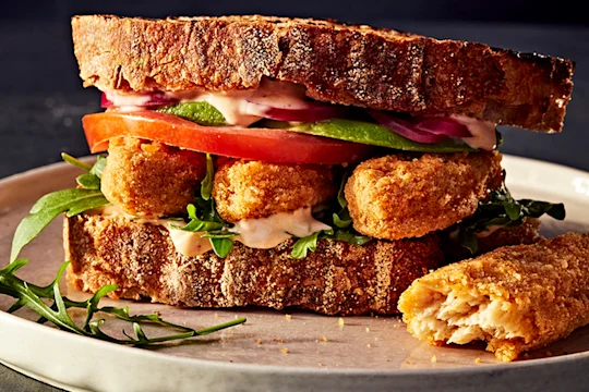 Crispy vegetarian chicken sandwich with tomato, onion and salad in sourdough bread served on a white plate with a cut Quorn Salt and Pepper Tender on the side.