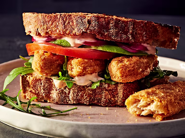 Crispy vegetarian chicken sandwich with tomato, onion and salad in sourdough bread served on a white plate with a cut Quorn Salt and Pepper Tender on the side.