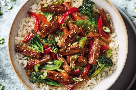 A Quorn Steak Strips stir-fry with carrots, broccoli, peppers, mange tout in a teriyaki sauce with rice in a white bowl on a blue background.