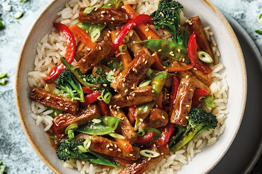 A Quorn Steak Strips stir-fry with carrots, broccoli, peppers, mange tout in a teriyaki sauce with rice in a white bowl on a blue background.
