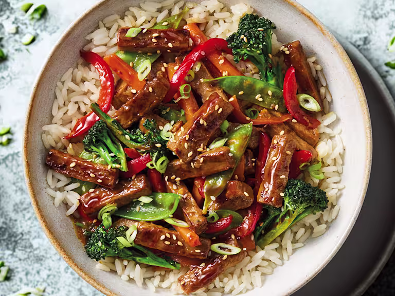 A Quorn Steak Strips stir-fry with carrots, broccoli, peppers, mange tout in a teriyaki sauce with rice in a white bowl on a blue background.