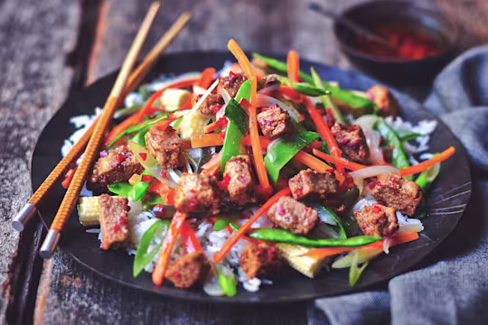 Quorn ChiQin Pieces stir-fried with pepper, carrots, snow peas, and broccolini atop rice on a black plate with a pair of chopsticks.