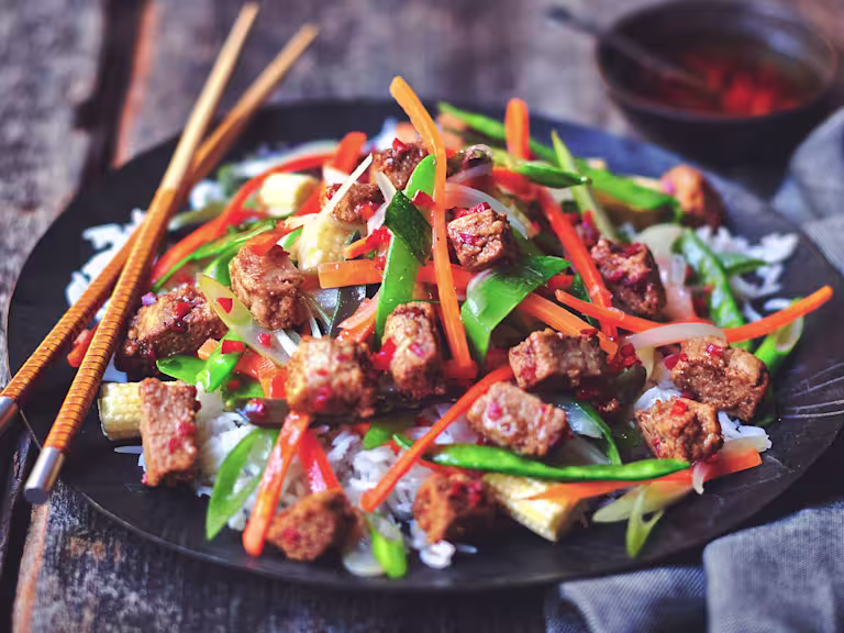 Quorn ChiQin Pieces stir-fried with pepper, carrots, snow peas, and broccolini atop rice on a black plate with a pair of chopsticks.