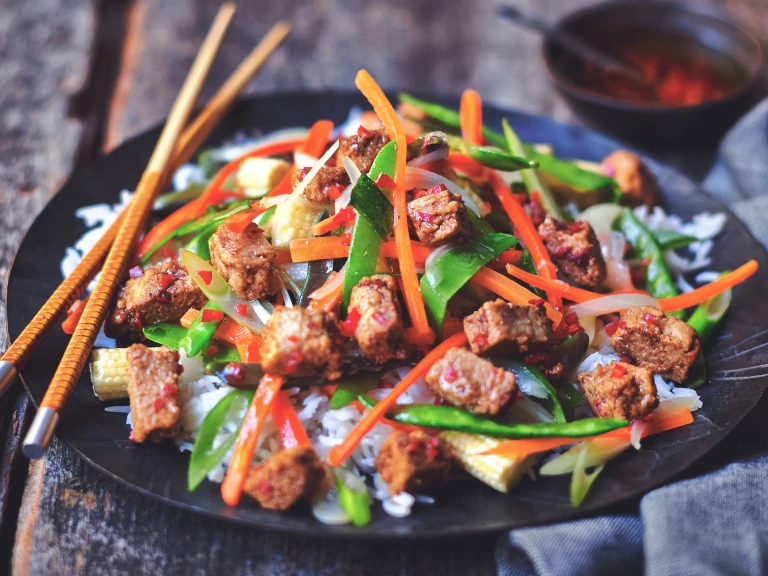 Quorn Pieces stir-fried with pepper, carrots, snow peas, and broccolini atop rice on a black plate with a pair of chopsticks.