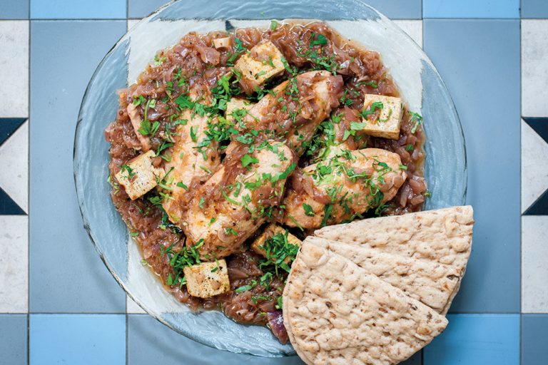 A glass bowl of doro wat made with Quorn Fillets on a blue tile background with two pieces of flatbread on the side.