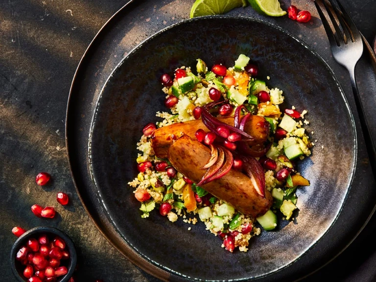 Two Quorn Sticky Glazed sausages topped with pomegranate seeds on a bed of pilaf cous-cous with a side serving of pomegranate seeds.