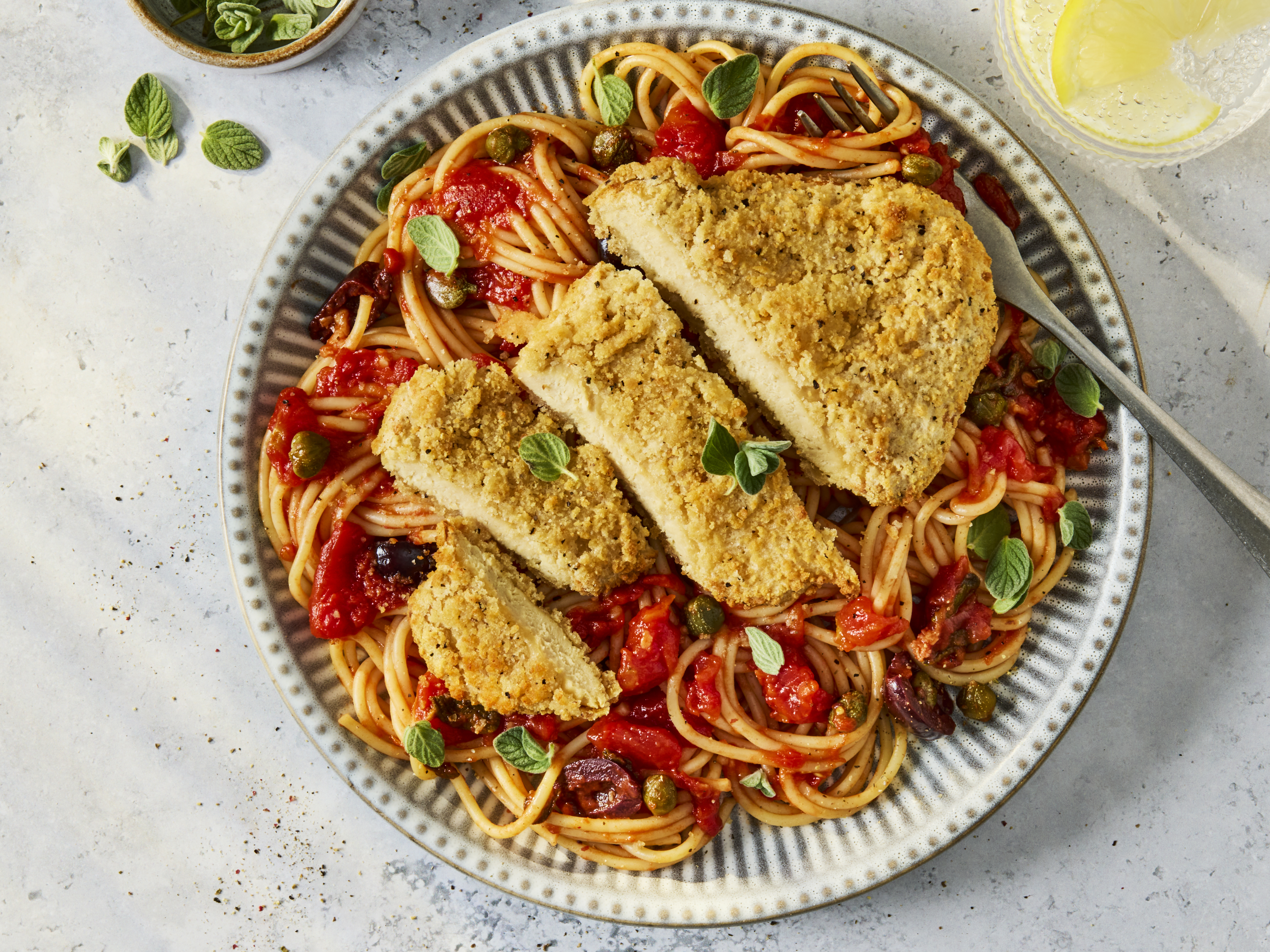 A bowl of vegetarian pasta puttanesca with rich tomato sauce, olives, and capers, topped with a sliced golden-brown Quorn Lemon and Pepper Schnitzel and garnished with fresh oregano leaves.