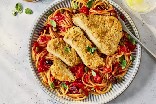 A bowl of vegetarian pasta puttanesca with rich tomato sauce, olives, and capers, topped with a sliced golden-brown Quorn Lemon and Pepper Schnitzel and garnished with fresh oregano leaves.