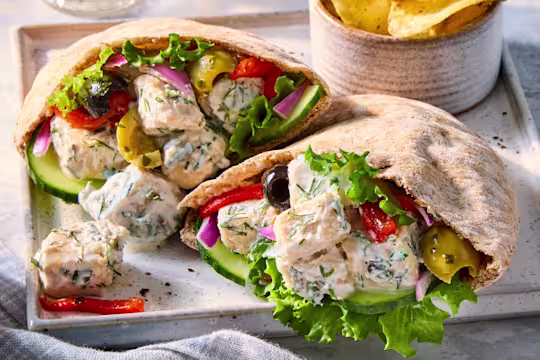 Two Greek-inspired Vegetarian Pitas with Quorn Pieces, filled with lettuce, cucumber, peppers and olives, on a grey serving tray next to a bowl of crisps.