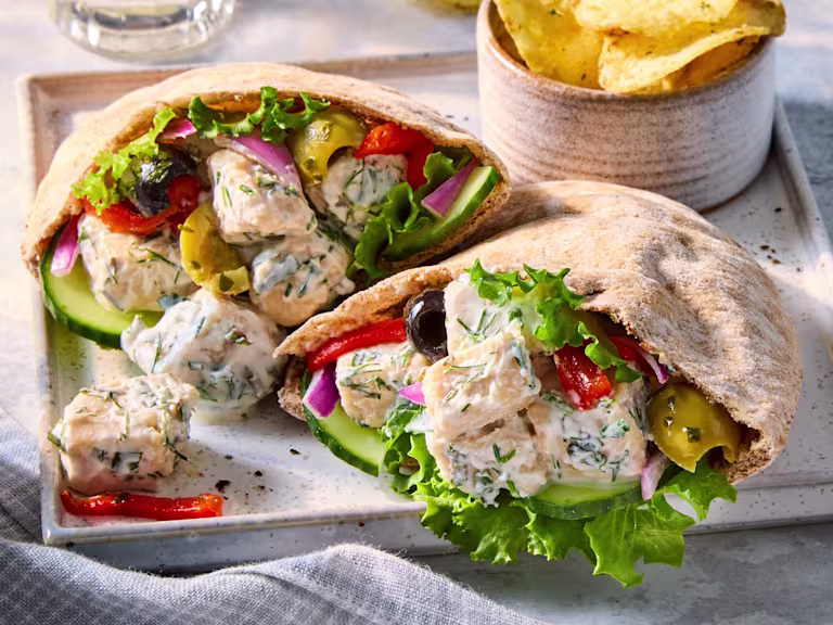 Two Greek-inspired Vegetarian Pitas with Quorn Pieces, filled with lettuce, cucumber, peppers and olives, on a grey serving tray next to a bowl of crisps.