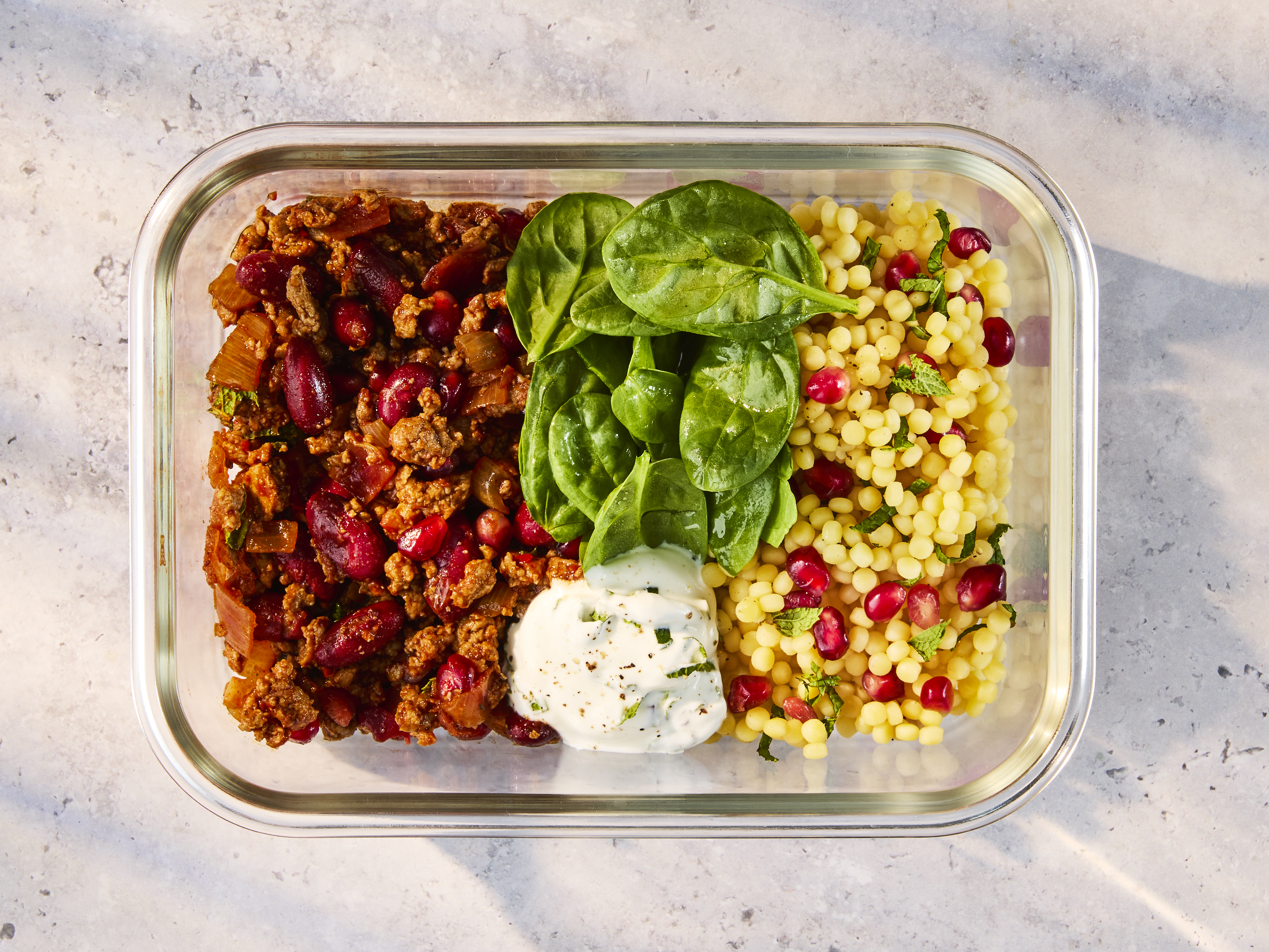 A glass meal prep container holding Quorn Persian Mince with Couscous, fresh spinach, and Mint Yoghurt.