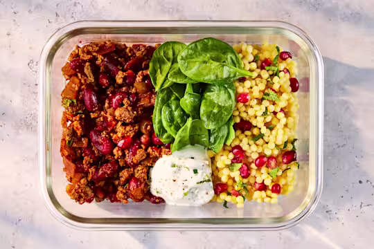 A glass meal prep container holding Quorn Persian Mince with Couscous, fresh spinach, and Mint Yoghurt.