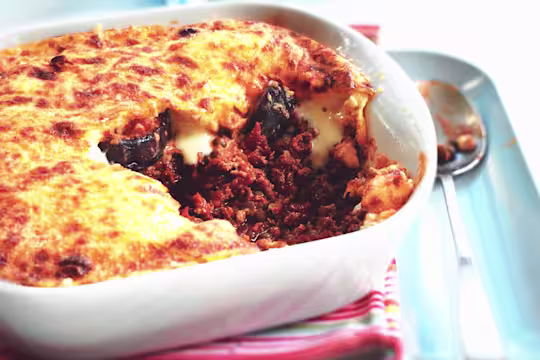 A baked Quorn moussaka in a white ceramic dish, with a portion removed to show layers of meat-free Quorn mince in a rich tomato sauce, slices of eggplant, and a golden, bubbling béchamel topping. A spoon with some filling rests nearby