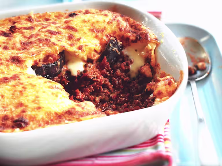 A baked Quorn moussaka in a white ceramic dish, with a portion removed to show layers of meat-free Quorn mince in a rich tomato sauce, slices of eggplant, and a golden, bubbling béchamel topping. A spoon with some filling rests nearby