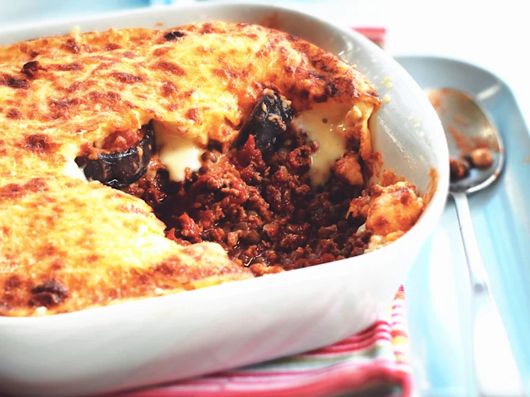 A baked Quorn moussaka in a white ceramic dish, with a portion removed to show layers of meat-free Quorn mince in a rich tomato sauce, slices of eggplant, and a golden, bubbling béchamel topping. A spoon with some filling rests nearby