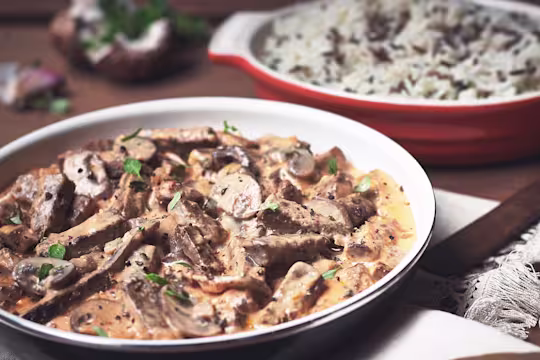 Close-up of a creamy Quorn Vegetarian Steak Strips and Mushroom Stroganoff served in a bowl, garnished with fresh herbs. A dish of rice is in the background, completing this comforting vegetarian meal.