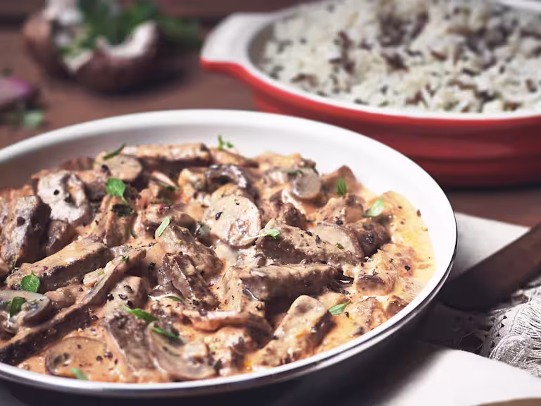 Close-up of a creamy Quorn Vegetarian Steak Strips and Mushroom Stroganoff served in a bowl, garnished with fresh herbs. A dish of rice is in the background, completing this comforting vegetarian meal.