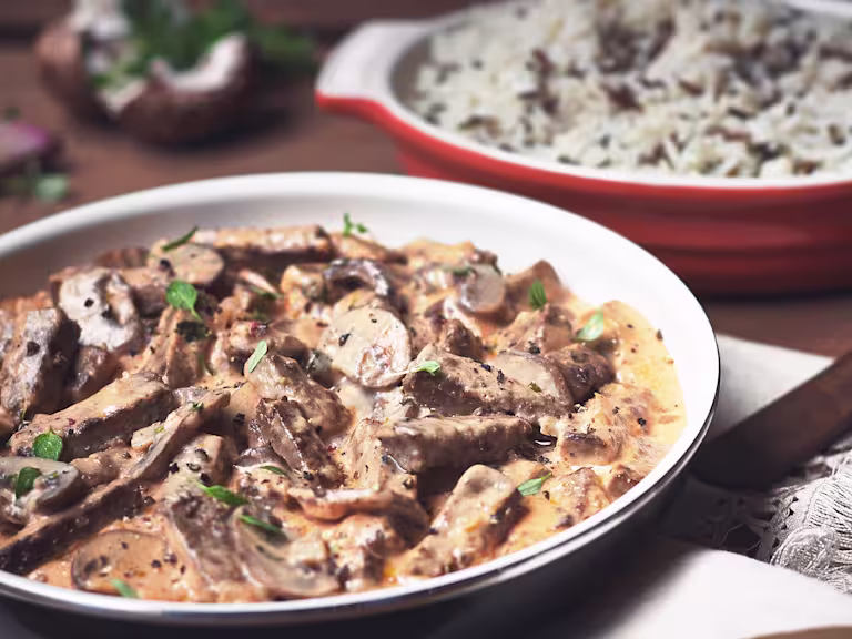Close-up of a creamy Quorn Vegetarian Steak Strips and Mushroom Stroganoff served in a bowl, garnished with fresh herbs. A dish of rice is in the background, completing this comforting vegetarian meal.