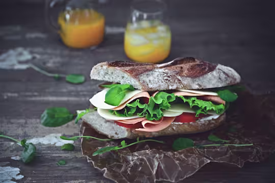A sandwich on a rustic baguette filled with Quorn Vegan Ham Slices, tomato slices, lettuce, and cheese with a carafe and glass of orange juice in the background.