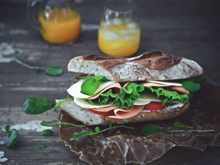 A sandwich on a rustic baguette filled with Quorn Vegan Ham Slices, tomato slices, lettuce, and cheese with a carafe and glass of orange juice in the background.