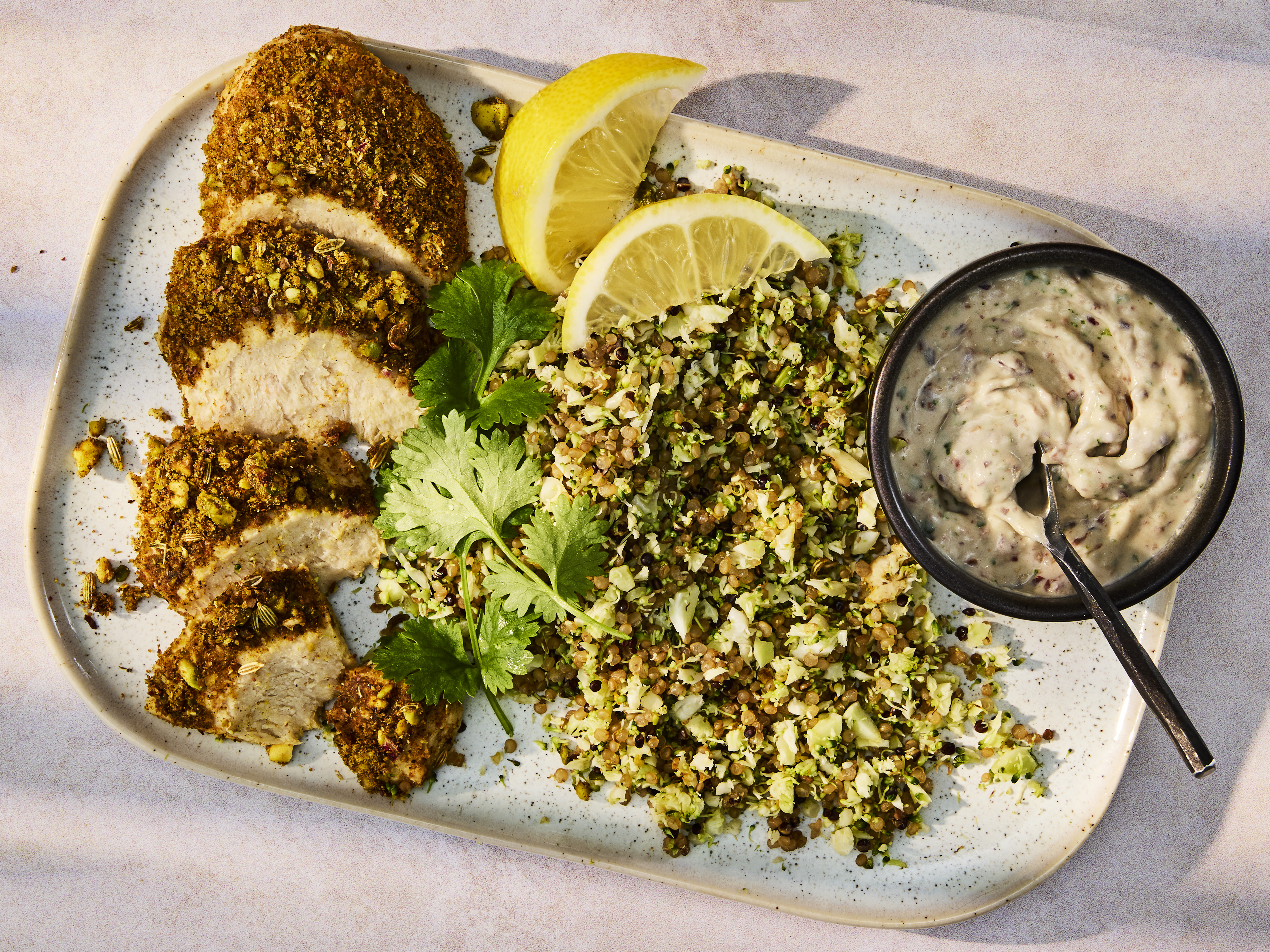 Sliced Dukkha Quorn Fillets on a speckled rectangular plate, served with a green herb and grain salad, lemon wedges, and a small dark bowl of creamy 5 Bean Hummus.