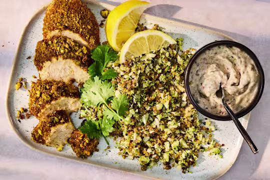 Sliced Dukkha Quorn Fillets on a speckled rectangular plate, served with a green herb and grain salad, lemon wedges, and a small dark bowl of creamy 5 Bean Hummus.