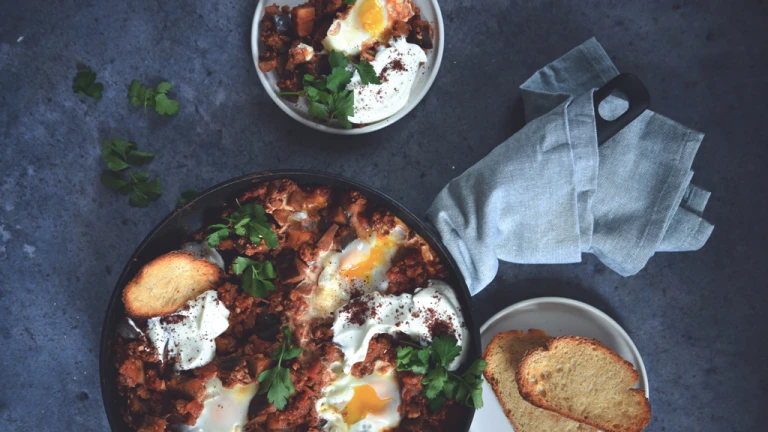 Shakshuka of Quorn Mince and aubergine and eggs, served in a dish next to a plate of toasts and a serving of shakshuka