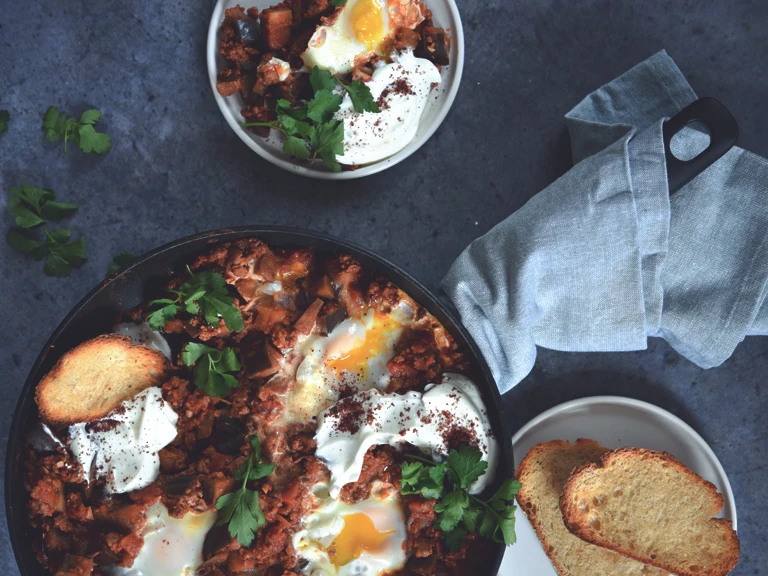 Shakshuka of Quorn Mince and aubergine and eggs, served in a dish next to a plate of toasts and a serving of shakshuka