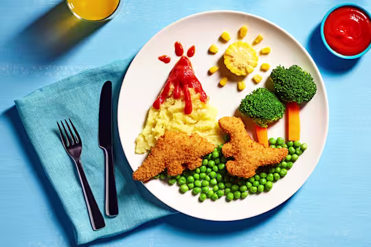 Two Quorn Roarsomes Vegan Dinosaur Nuggets against a backdrop of peas, carrot and broccoli trees, and a mashed potato volcano on a white plate.