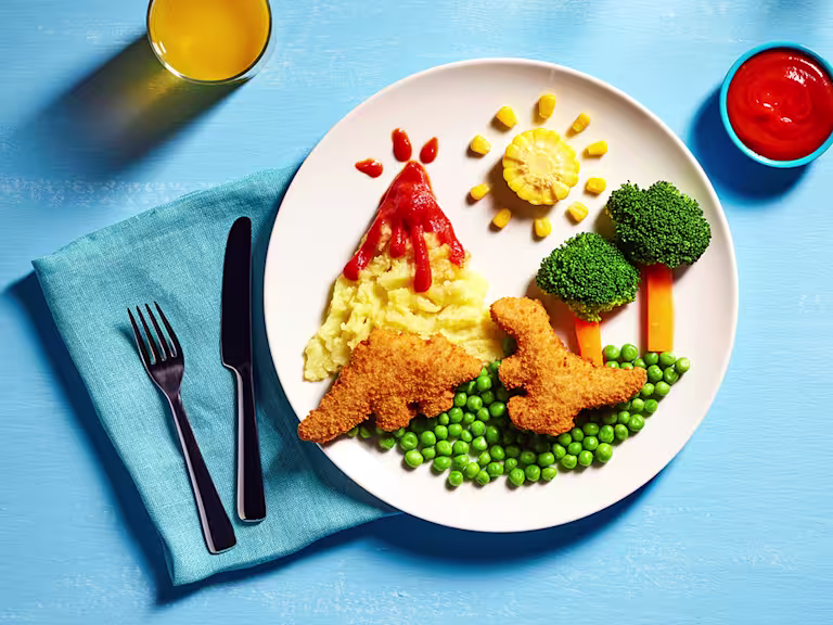 Two Quorn Roarsomes Vegan Dinosaur Nuggets against a backdrop of peas, carrot and broccoli trees, and a mashed potato volcano on a white plate.