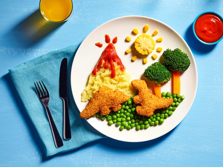 Two Quorn Roarsomes Vegan Dinosaur Nuggets against a backdrop of peas, carrot and broccoli trees, and a mashed potato volcano on a white plate.
