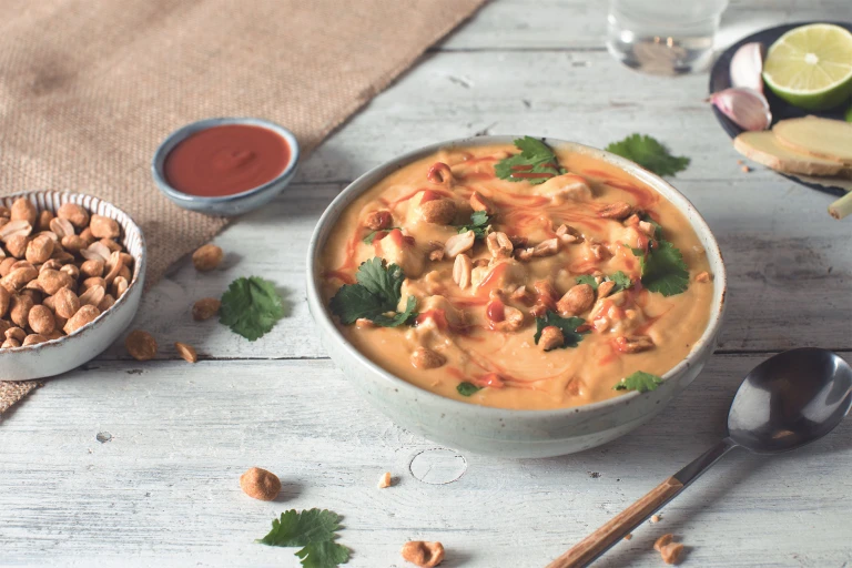 A bowl of soup topped with Quorn Pieces, cilantro, peanuts, and sriracha with smaller bowls of peanuts and sriracha in the backgrounds.