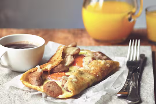 A galette filled with Quorn Sausage, cheese, and two eggs on a piece of paper with a cup of coffee and cutlery on either side.