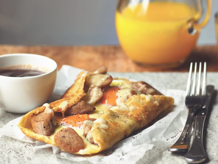 A galette filled with Quorn Sausage, cheese, and two eggs on a piece of paper with a cup of coffee and cutlery on either side.