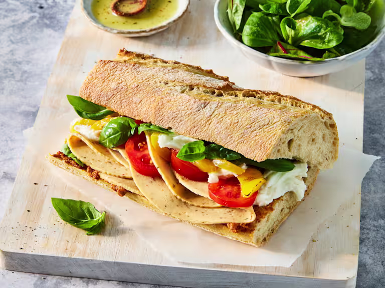 White baguette with Quorn Tomato & Basil Slices, mozzarella, roasted yellow peppers and fresh tomato and basil on a chopping board, with dressing and spinach on the side.