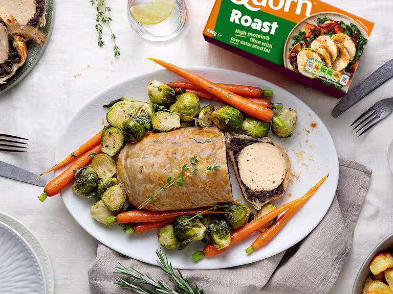 A vegetarian wellington made with Quorn Roast wrapped in mushrooms duxelles and puff pastry surrounded by roasted brussels sprouts and carrots with roast potatoes on the side.