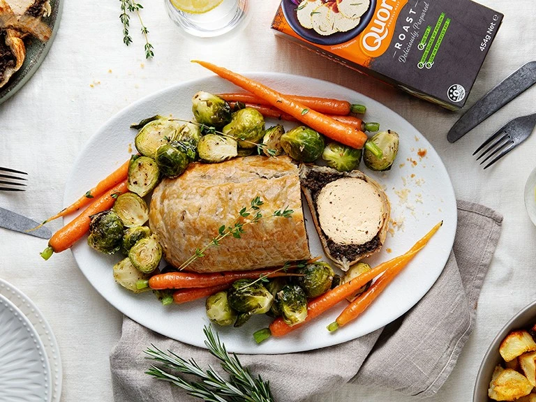 A vegetarian wellington made with Quorn Roast wrapped in mushrooms duxelles and puff pastry surrounded by roasted brussels sprouts and carrots with roast potatoes on the side.