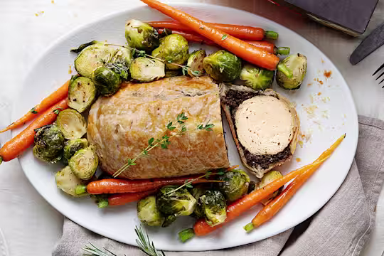 A vegetarian wellington made with Quorn Roast wrapped in mushrooms duxelles and puff pastry surrounded by roasted brussels sprouts and carrots with roast potatoes on the side.