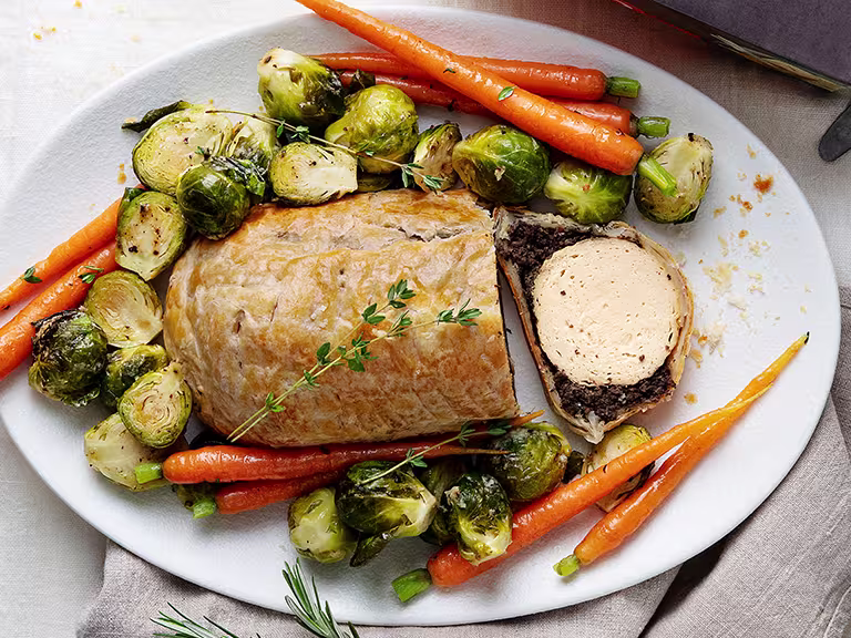 A vegetarian wellington made with Quorn Roast wrapped in mushrooms duxelles and puff pastry surrounded by roasted brussels sprouts and carrots with roast potatoes on the side.