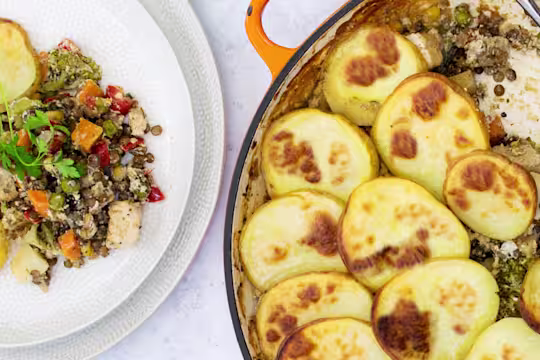A lentil hotpot with Quorn Meat Free Pieces and mixed vegetables topped with sliced potatoes in a shallow orange Dutch oven with one portion removed and plated on the side.