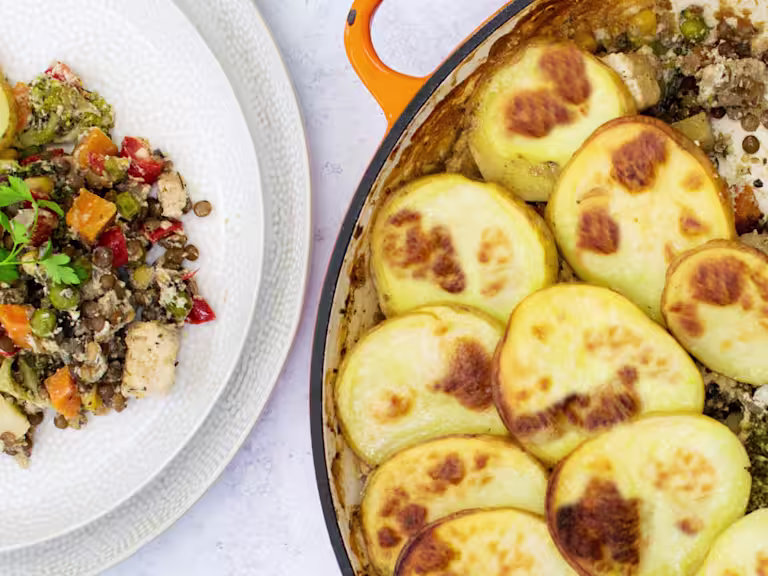 A lentil hotpot with Quorn Meat Free Pieces and mixed vegetables topped with sliced potatoes in a shallow orange Dutch oven with one portion removed and plated on the side.